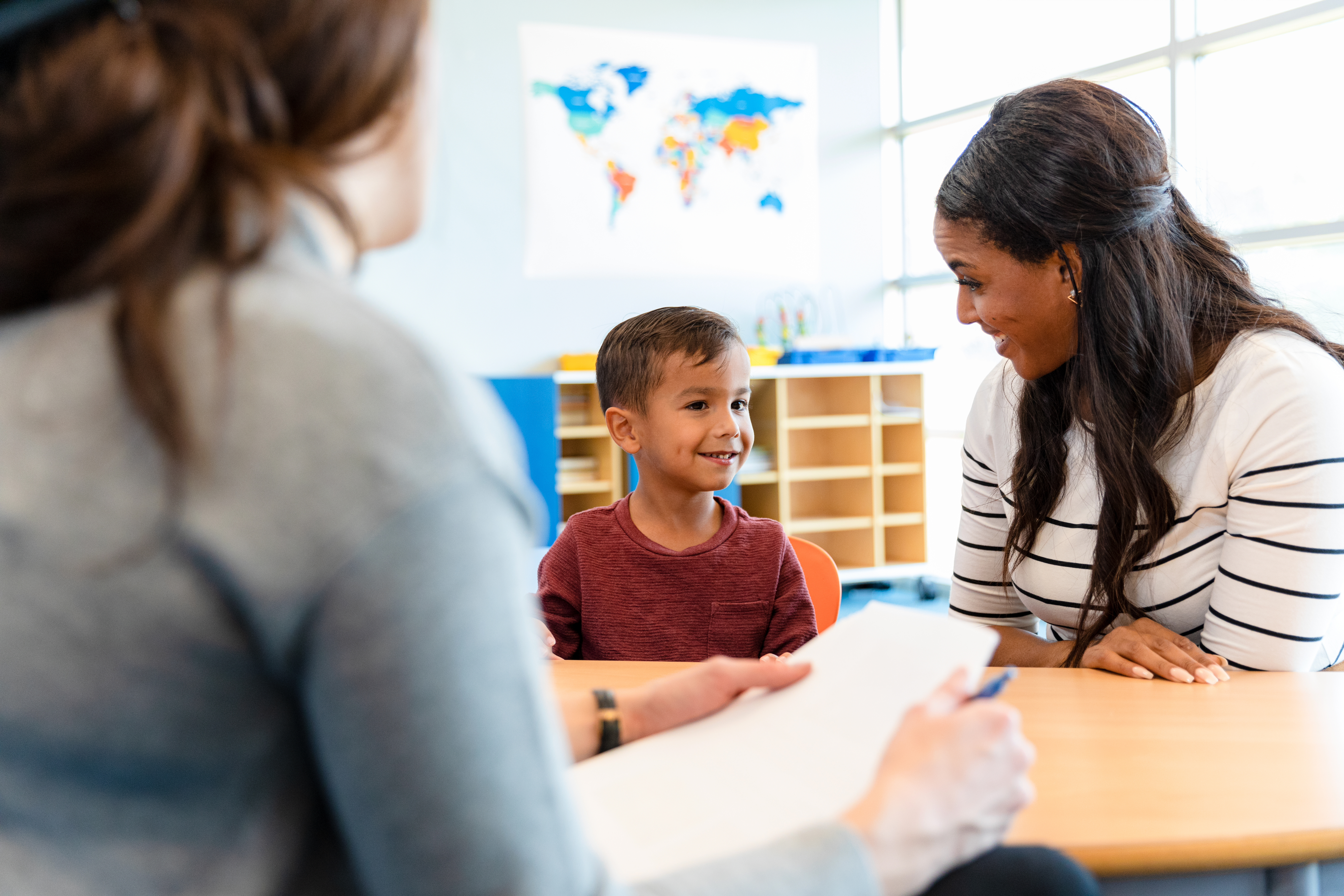 A small boy wearing a burgundy t-shirt sits at a table smiling with a woman wearing a striped top sitting next to him and smiling, looking at the boy. Another woman wearing a long sleeved, grey top, sits at the opposite end of the table with her back to the camera.