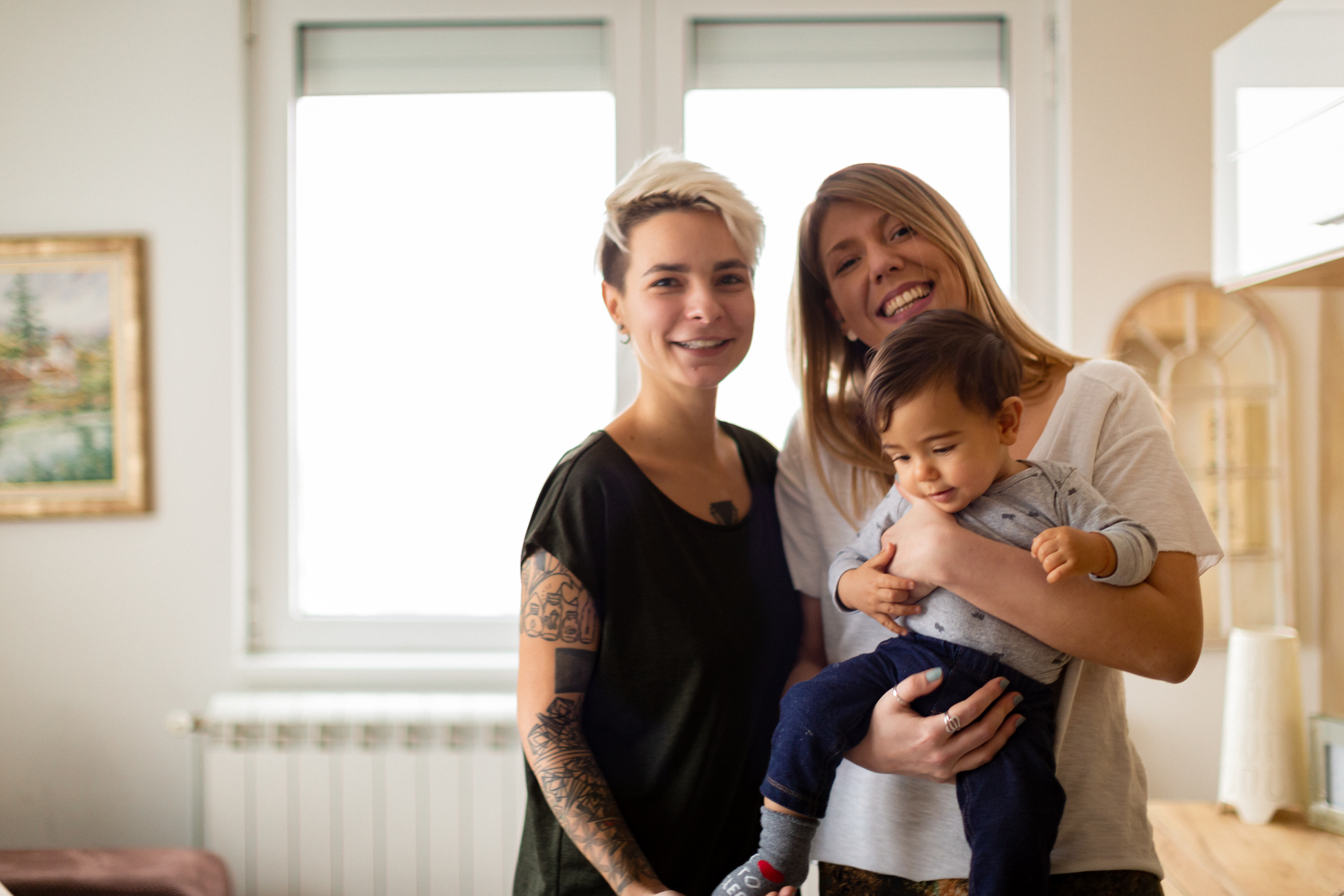 Two women stand together, smiling, one is wearing a black t-shirt, the other is wearing a white t-shirt. The woman in the white t-shirt is holding a baby boy, about 12 months old. in a long sleeved grey top and navy pants.