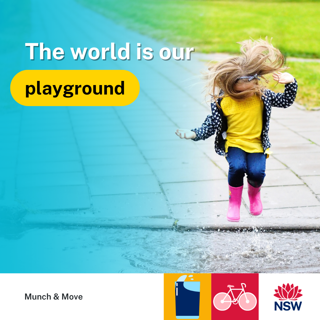 young girl with long blond hair obscuring her face, wearing a yellow shirt, blue jacket and pink gumboots. Jumping in a puddle on a path making a splash